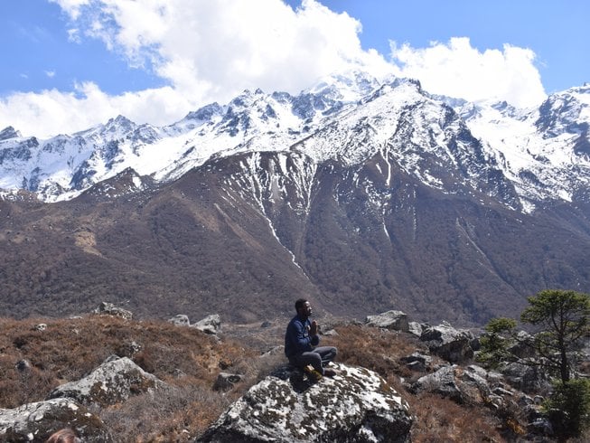 A solo yogi sits in an introspective, cross-legged meditation pose with hands in prayer atop a large boulder, facing a towering, jagged alpine mountain wall streaked with deep winter snow.