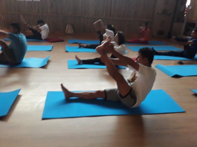A class of practitioners spread out across blue mats on a smooth wooden floor, performing an active seated leg stretching sequence inside a spacious, rustic yoga shala.