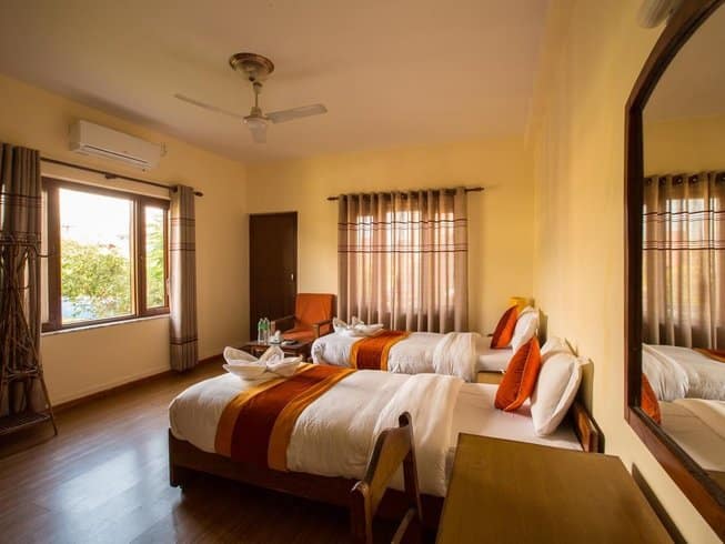 A bright and warm twin-sharing bedroom featuring modern wood floors, two clean beds with white linens and orange runners, and a large mirror reflecting warm afternoon sunlight streaming in from the window.