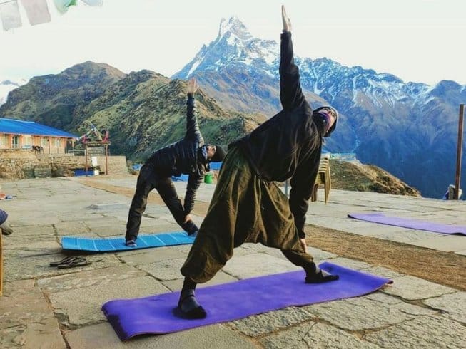 A male and female practitioner hold dynamic and grounded triangle yoga poses on purple mats, set against the breathtaking vertical relief of a sharp, snow-capped mountain peak (possibly Machhapuchhre or Fishtail) and lush green valley ridges.