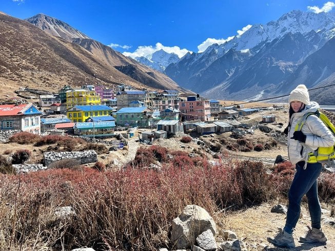 A female trekker with a bright yellow backpack and white knit beanie poses on a dirt trail overlooking a picturesque mountain valley village filled with colorful yellow, blue, and pink teahouses.