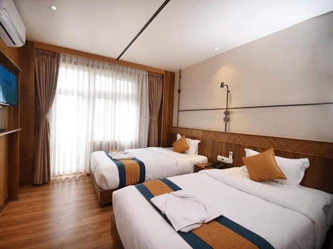 A warm, modern twin-sharing lodge bedroom in Nepal featuring premium hardwood flooring, crisp white bedding with blue and gold runners, and a minimalist pendant reading light over the headboard.