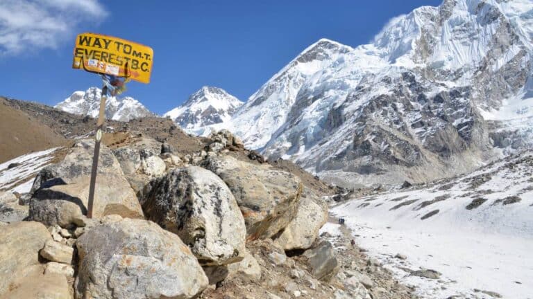 A rustic yellow wooden trail sign reading 'WAY TO M.T. EVEREST B.C.' stands on a rocky, snow-dusted ridge, overlooking a vast mountain valley with massive snow-capped peaks under a bright blue sky.