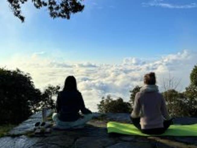 Rear silhouette view of two women meditating cross-legged on yoga mats atop a stone viewpoint, looking out over a breathtaking, endless sea of thick white clouds stretching toward a blue horizon.