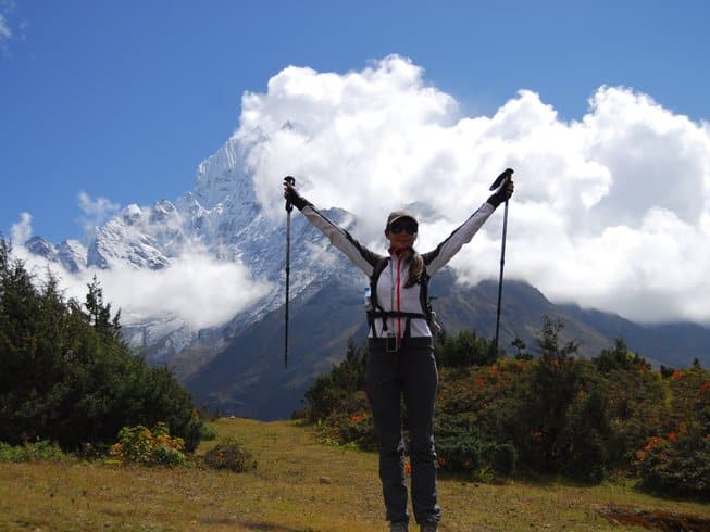 A wide-angle landscape shot of a female hiker with gray pants, a white and black jacket, and a baseball cap standing on a grassy path with her arms raised, holding hiking poles triumphantly. In the background is a large, sharp, snow-covered mountain peak, partially obscured by clouds, with green juniper bushes scattered in the foreground.